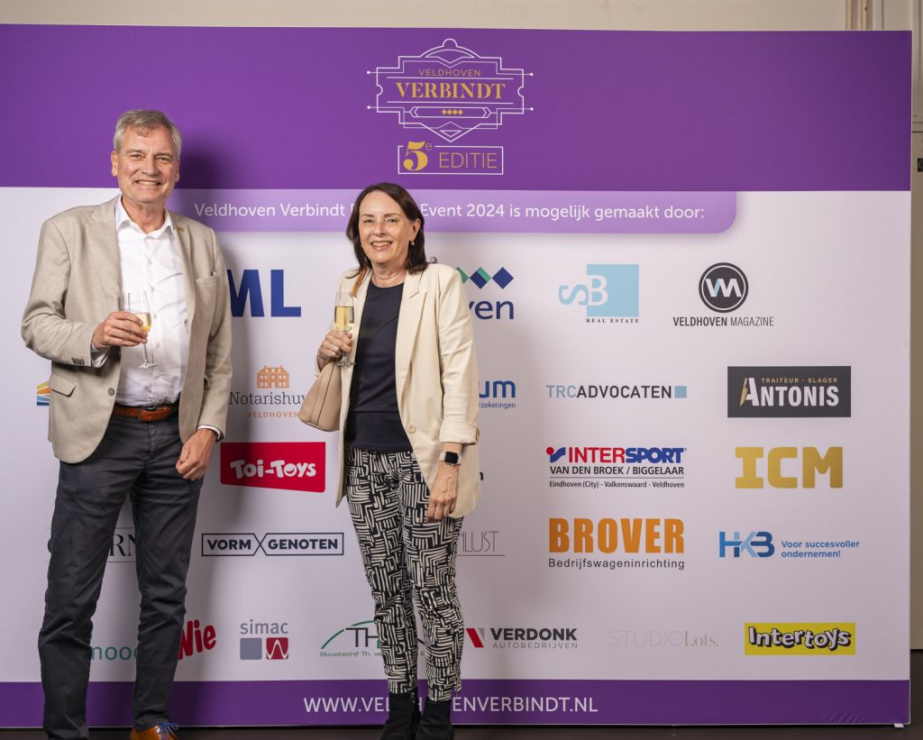 Twee mensen met champagneglazen bij sponsorsbord op evenement.