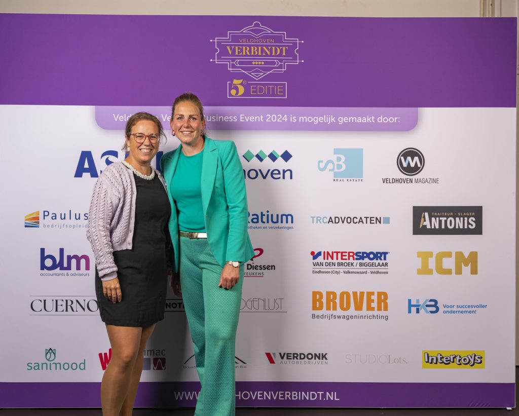Twee vrouwen bij sponsorbord op zakelijk evenement.