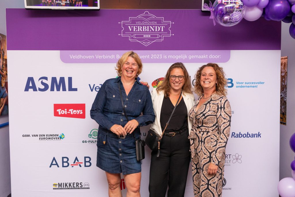 Drie vrouwen poseren voor sponsorbord evenement.