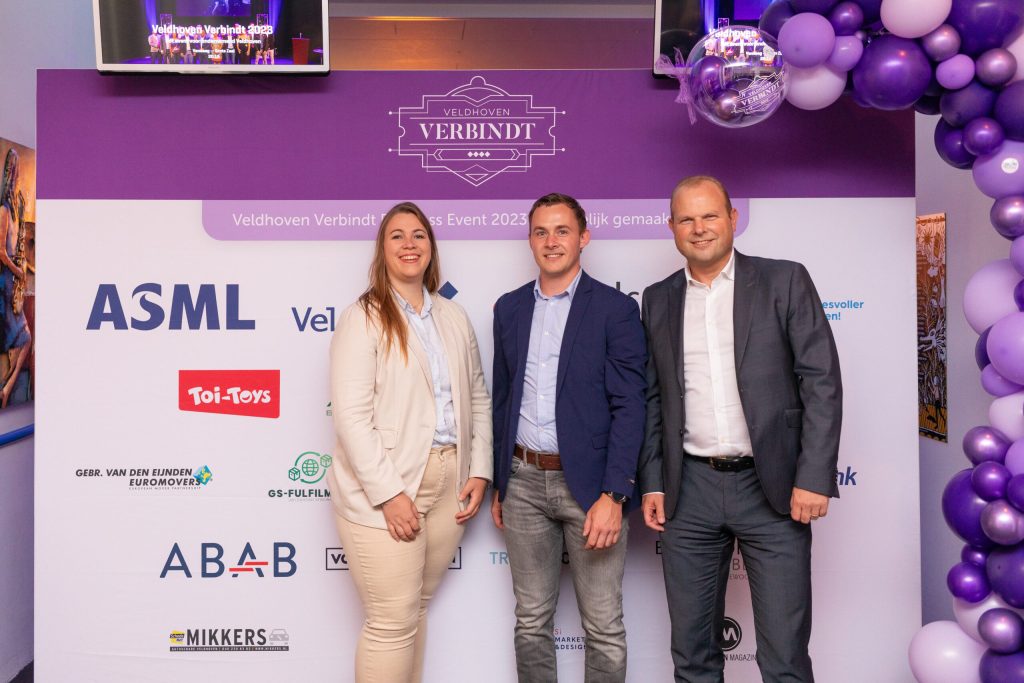 Drie mensen bij Veldhoven Verbindt bord met logo's.