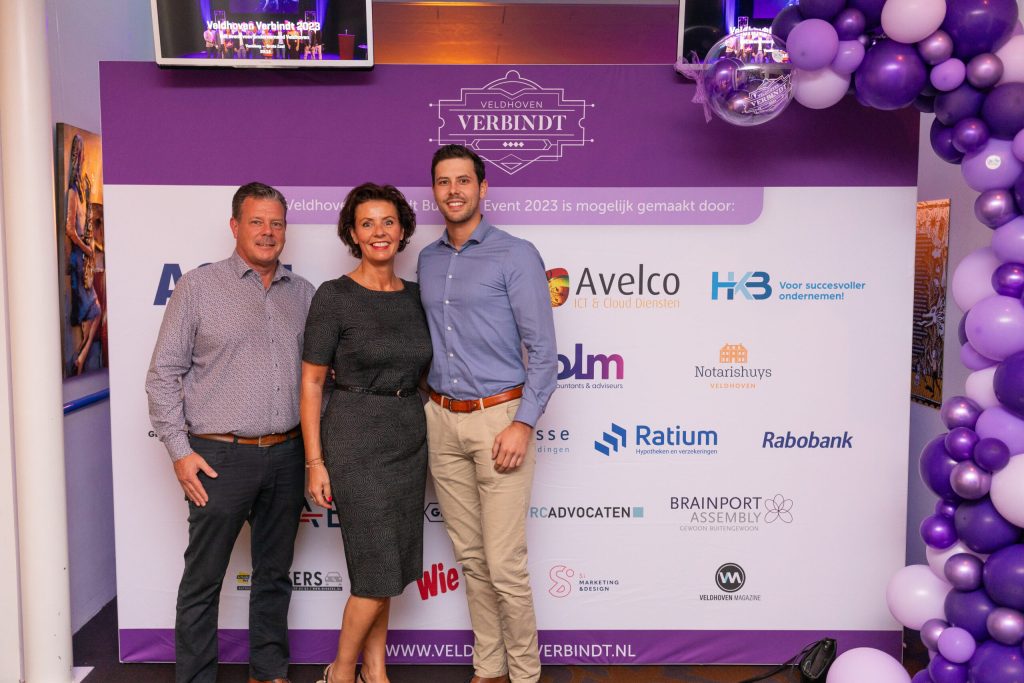 Drie mensen poseren voor Veldhoven Verbindt sponsorbord.